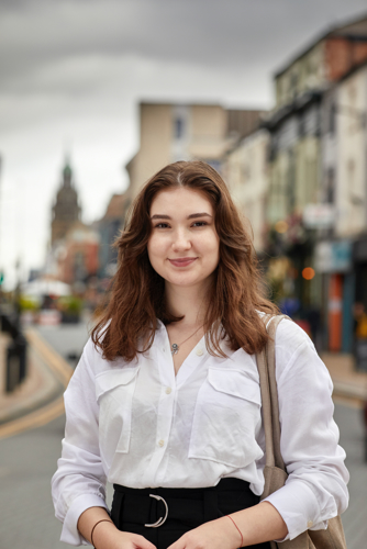 A person wearing a white button-up shirt and black belted trousers is standing on a city street with buildings lining both sides. The individual has a beige shoulder bag and is positioned in the foreground, while the background shows a slightly overcast sky and a distant clock tower. The street features double yellow lines and appears to be in an urban area with shops and pedestrians.