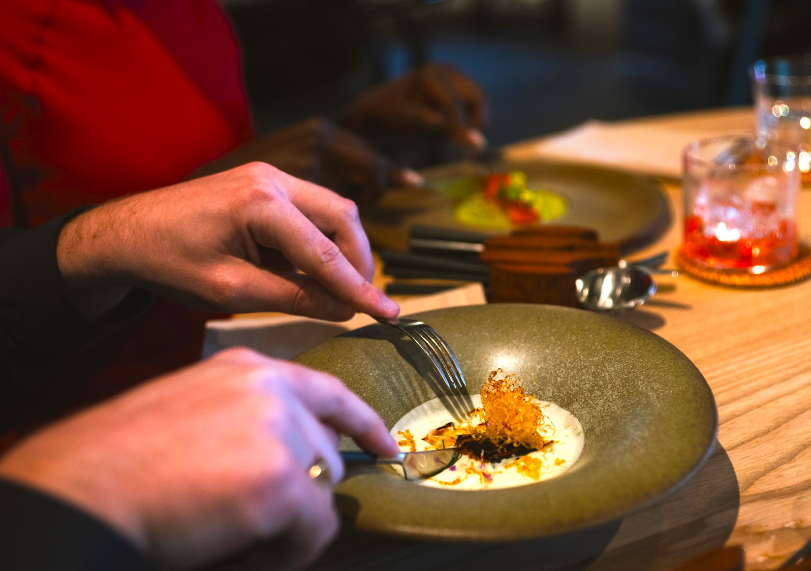 Close‑up of diners enjoying a plated fine‑dining dish at a restaurant table, with food being eaten using cutlery.