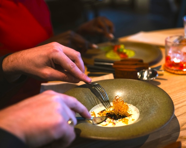 Close‑up of diners enjoying a plated fine‑dining dish at a restaurant table, with food being eaten using cutlery.