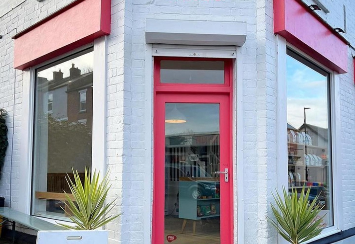 A brick building that has been painted white, with red window frames and door. There is a hand painted sign, in red, above the door that reads 'Corner Store'. The letter 'o' in the word Store is a smiley face.