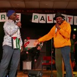 :Two performers stand on a stage holding microphones during a live event. One wears a patterned shirt with a Palestinian scarf and blue hat, while the other wears a bright orange hoodie and black hat. Behind them, a banner spells “FREE PALESTINE” in large letters. Musical equipment, a chair and stage crew are visible in the background at The Crookes Working Mens Club. Image Credit: Leon Lockley.