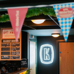 Interior of a beer hall featuring a bar counter with a chalkboard-style menu listing guest beers and prices. A neon sign with the letter “K” is illuminated on a dark wall near a wooden door. Colourful triangular bunting hangs across the ceiling, and green garland decorates the space. The setting has warm lighting and a modern, welcoming atmosphere.