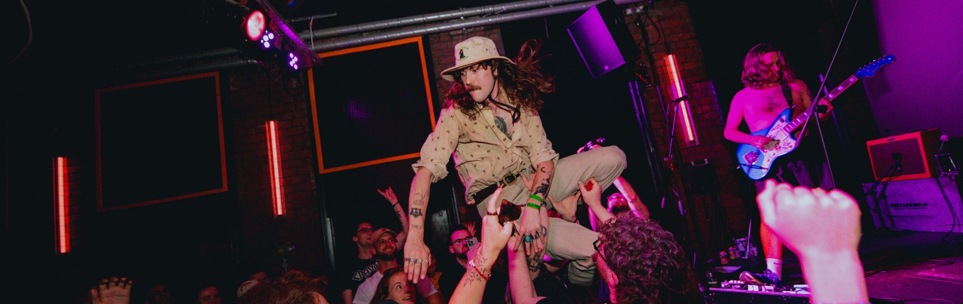 A lively concert scene in a dimly lit venue, with a performer being lifted by the crowd during a stage dive. The audience is packed closely together, raising their hands to support the performer. Bright pink and purple stage lights illuminate the space, and a guitarist plays in the background near amplifiers. The ceiling features bold black-and-white curved patterns, adding to the energetic atmosphere.