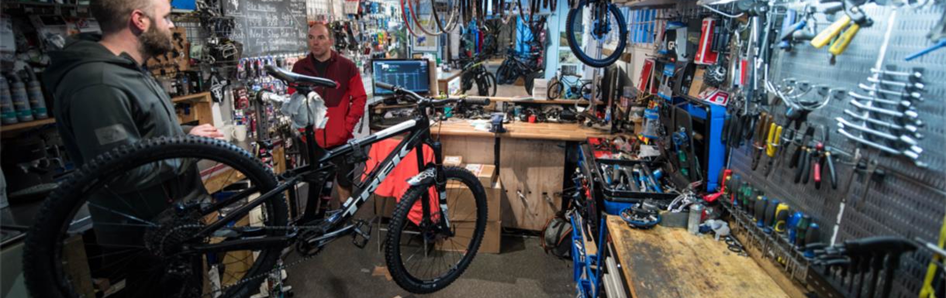 A photograph of the interior of The Bike Garage with the walls covered in tools and spare parts. There are two men working on bikes.