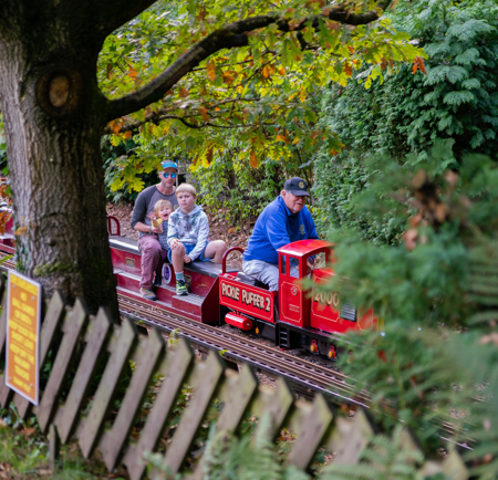 A small red miniature train travels along a track surrounded by trees and greenery. Several passengers are seated on open carriages behind the engine, passing a wooden fence and a yellow sign in a leafy outdoor setting.