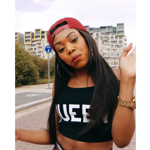 Portrait photo of a woman standing outdoors on a street with modern apartment buildings in the background. She has long dark hair, wears a red baseball cap backwards, large hoop earrings, a black crop top with white lettering, and a gold watch. She tilts her head and raises one hand in a playful, expressive gesture while looking towards the camera.