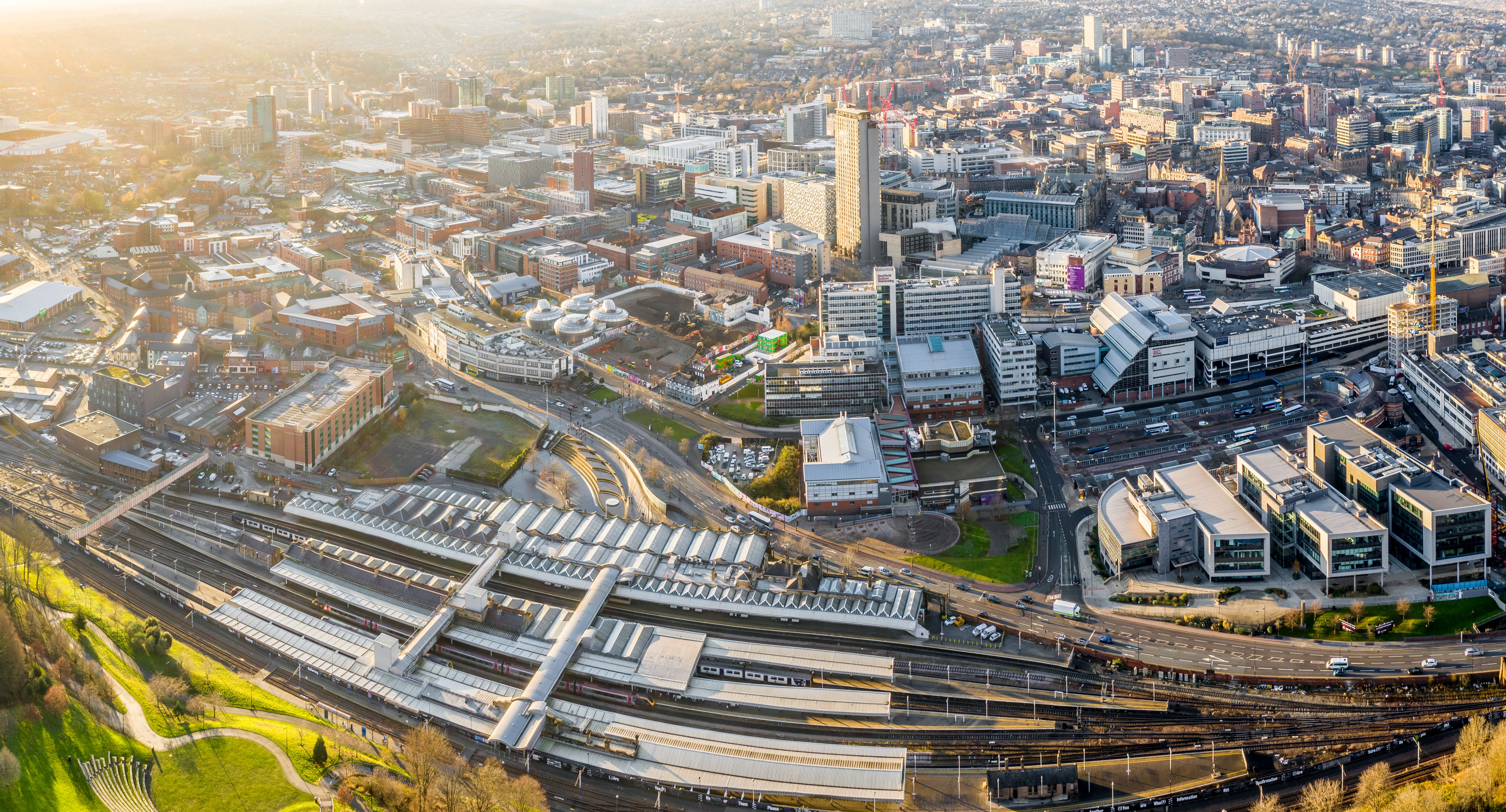 Aerial view of Sheffield city centre in warm sunlight, showing the railway station in the foreground with multiple platforms and tracks, surrounded by modern office buildings, roads, and ongoing construction sites leading into the wider urban landscape.