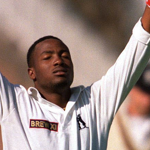 A cricketer in a white long-sleeved shirt celebrates on the field with both arms raised high. Red wristbands are visible on each wrist, and a blurred crowd and players form the background. The close-up image captures a moment of triumph and achievement during a cricket match, with the athlete’s expression conveying pride and determination.