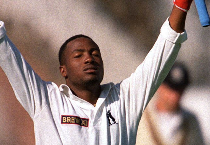 A cricketer in a white long-sleeved shirt celebrates on the field with both arms raised high. Red wristbands are visible on each wrist, and a blurred crowd and players form the background. The close-up image captures a moment of triumph and achievement during a cricket match, with the athlete’s expression conveying pride and determination.