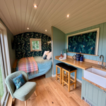 Compact and stylish cabin interior with pale blue panelled walls, a curved white ceiling and light wood flooring. A double bed with soft blue bedding and a colourful woven throw sits beneath dark botanical wallpaper featuring leafy branches and birds. Opposite, a small kitchenette includes a wooden worktop, Belfast sink and two bar stools. A cushioned chair stands by a large window, filling the cosy space with natural light and views of greenery outside.