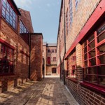  A shot of Leah's Yard, in the sunshine, in the centre of Sheffield. A cobbled path is lined on both sides by renovated red brick industrial buildings.