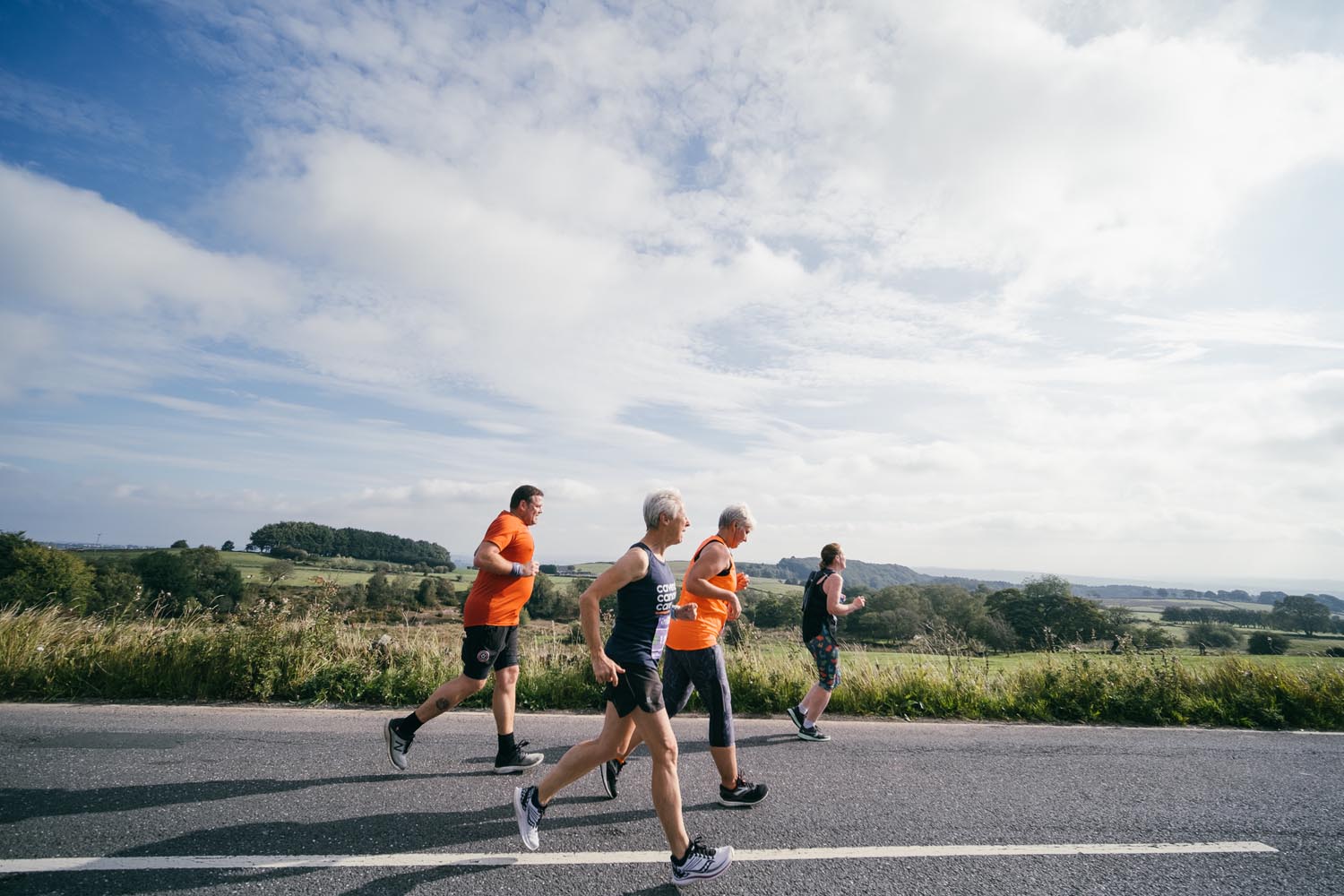 Four people jogging along a rural road on a bright day, with open countryside and green fields stretching into the distance under a partly cloudy sky.