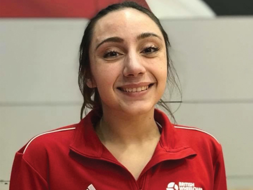 Person wearing a red sports jacket with white stripes on the shoulders and the British Weight Lifting logo on the chest, standing indoors against a light-coloured wall with a red and black background element.