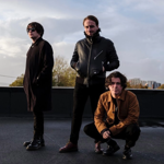 Three people pose on a rooftop or outdoor terrace under a cloudy sky. One stands wearing sunglasses and a long dark coat, another stands in a black leather jacket with hands in pockets, and a third crouches in a brown jacket and dark trousers. Buildings and trees are visible in the distance beyond a low wall. The image has a moody, urban promotional style, with soft evening light reflecting off the wet surface beneath them.