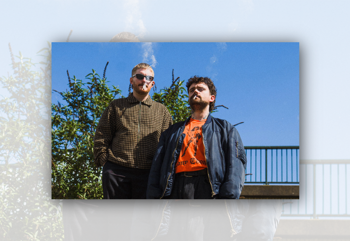 Two people stand outdoors beneath a clear blue sky, framed by leafy shrubs and a metal railing in the background. One wears sunglasses and a brown checked jacket, while the other wears a dark bomber jacket over a bright orange graphic T-shirt. Both are smoking cigarettes. The low-angle photograph creates a bold, stylised promotional portrait with strong sunlight and contrasting colours.