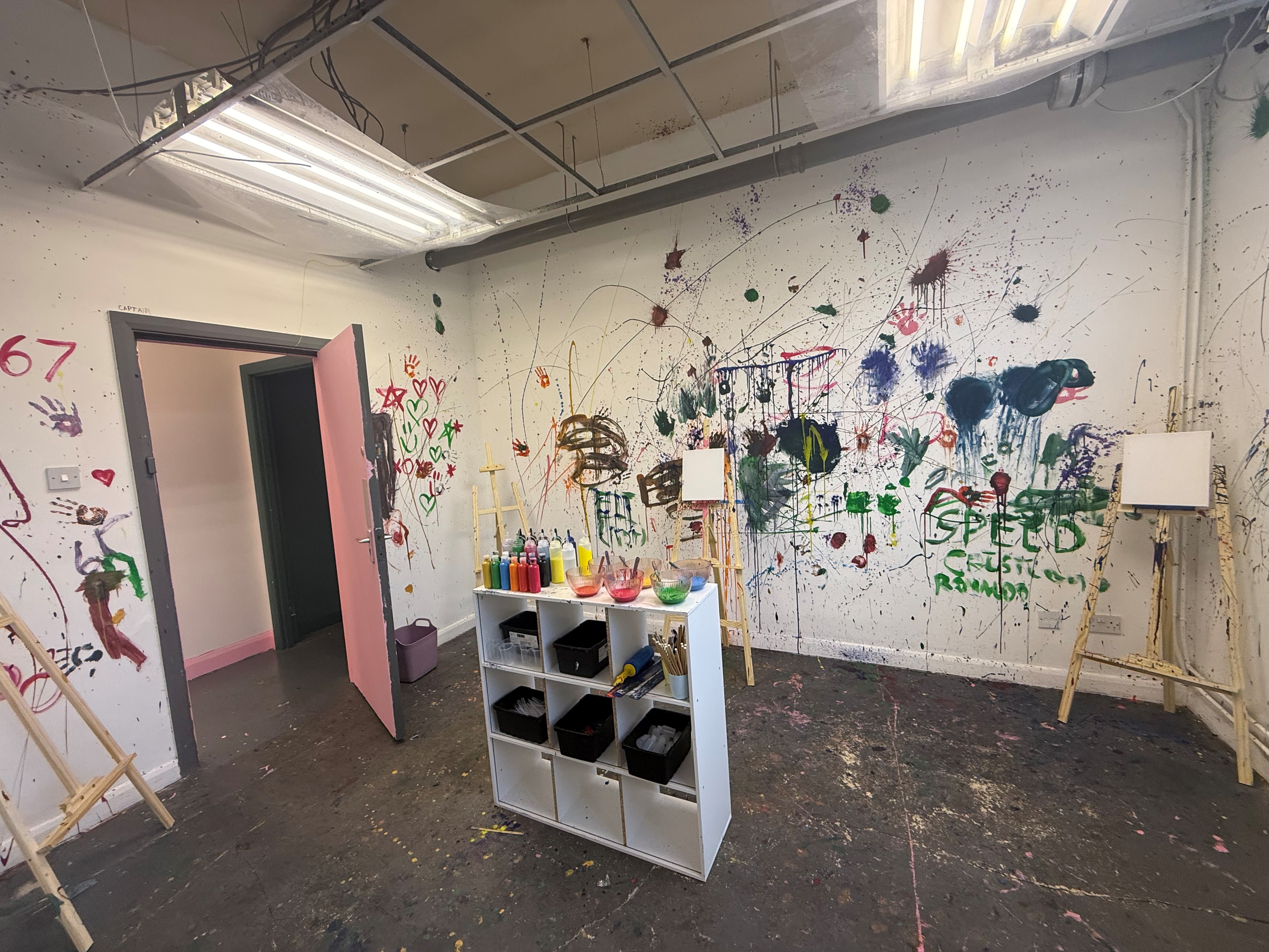 A paint-splattered art room with easels, colourful paint bottles and brushes, and walls covered in expressive splashes, doodles and handprints.
