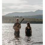Two people stand waist-deep in a lake, surrounded by shimmering water, with mountains and woodland in the background. One person holds an acoustic guitar while the other stands nearby in dark clothing. The scene is backlit by bright sunlight reflecting across the water, creating a soft, atmospheric landscape with distant hills beneath a cloudy sky.