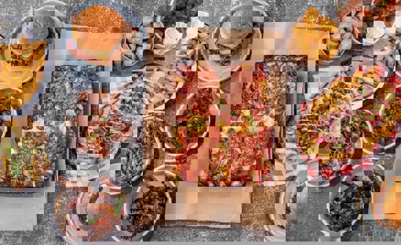 Overhead view of a food spread on a textured gray surface featuring a rectangular deep-dish pizza topped with pepperoni and cheese in the center. Surrounding dishes include burgers in buns, golden fries, crispy chicken wings, loaded fries with toppings, fried bites in a bowl, and dipping sauces. The arrangement showcases a variety of hearty comfort foods with vibrant colors and textures.