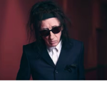 Studio-style portrait of a person with long dark hair wearing dark sunglasses, a dark tailored suit jacket and a white collared shirt. The person stands indoors against a softly lit background in deep red and burgundy tones. The image is closely cropped from the waist up, with warm lighting creating contrast between the dark clothing and the rich-coloured surroundings.