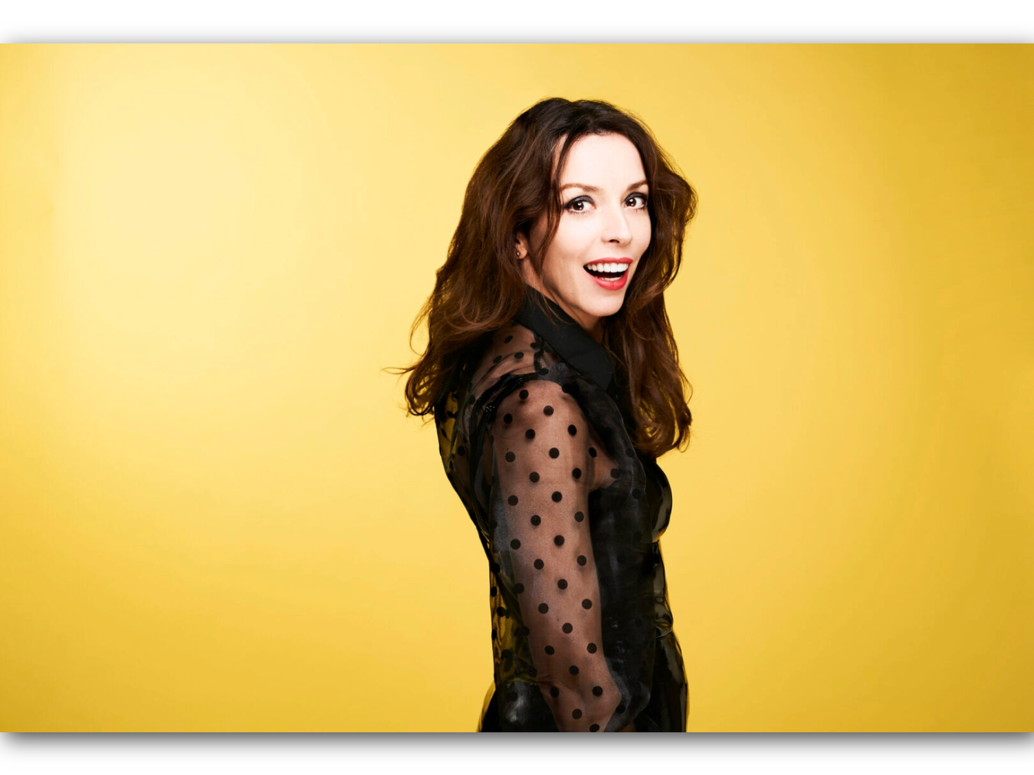Person standing against a bright yellow background, wearing a sheer black top with polka-dot sleeves and solid black panels. The individual has long, wavy brown hair and is posed in profile, facing slightly away from the camera.