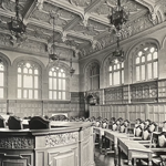 Black-and-white photograph of an ornate council chamber or assembly hall. Rows of carved wooden desks and chairs face a large central table with detailed decorative panels. Tall arched windows line the walls, filling the room with light. The high vaulted ceiling is richly decorated with intricate plasterwork, relief sculptures and hanging lantern-style chandeliers. Wood panelling surrounds the room, and a clock is mounted on the left wall. No people are present.