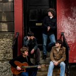Four musicians gather on the steps outside a building with a black doorway and red-painted wall. One person sits holding an acoustic guitar while three others sit or stand on the steps nearby. The scene is framed by textured stonework on one side and weathered red paint on the other, creating an informal band portrait in an urban setting.