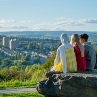 Three people sitting on a stone outcrop overlooking Sheffield, with terraced housing, green hills and open sky stretching across the city landscape.