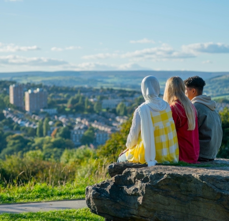 Three people sitting on a stone outcrop overlooking Sheffield, with terraced housing, green hills and open sky stretching across the city landscape.