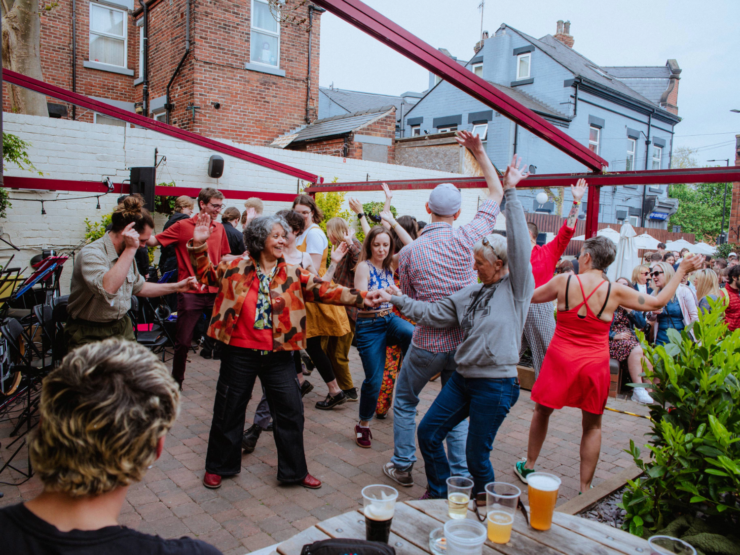 Outdoor gathering in a courtyard with people dancing and socializing under red beams. The space has brick flooring, wooden tables with drinks in the foreground, and a mix of greenery and brick buildings in the background. Some attendees are seated while others raise their arms and move energetically.