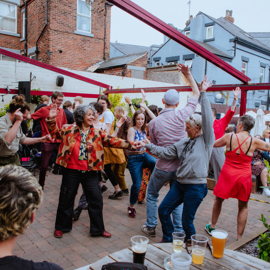 Outdoor gathering in a courtyard with people dancing and socializing under red beams. The space has brick flooring, wooden tables with drinks in the foreground, and a mix of greenery and brick buildings in the background. Some attendees are seated while others raise their arms and move energetically.