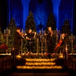 A string quartet performs on a stage, surrounded by many candles.