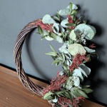 A half-covered wreath featuring a twisted vine base adorned with clusters of dried eucalyptus leaves, red foliage, and small white flowers. The arrangement is displayed on a wooden surface against a dark grey wall, showcasing an organic and asymmetrical design.