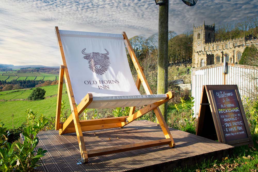 A large wooden deckchair printed with the Old Horns Inn logo stands on a raised platform overlooking rolling green countryside, with a historic stone church visible in the background under a cloudy sky.