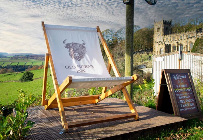 A large wooden deckchair printed with the Old Horns Inn logo stands on a raised platform overlooking rolling green countryside, with a historic stone church visible in the background under a cloudy sky.