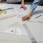 People working on pattern drafting at a large cutting table covered with pattern paper marked with measurements and guide lines. Hands hold paperwork and position a flexible measuring tape across the pattern. Drafting tools including clear rulers, a set square, curved ruler, pencil and tape measure are spread across the table, suggesting a garment design or sewing workshop.
