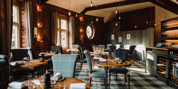 An elegant restaurant interior featuring round wooden tables set with folded napkins, glassware, and small decorative items. The space has exposed brick walls, large windows with gray curtains, and a patterned carpet. Pendant lights hang from the ceiling beams, creating warm ambient lighting. A wine storage area and shelving with glasses are visible on the right side of the room, adding to the sophisticated atmosphere.