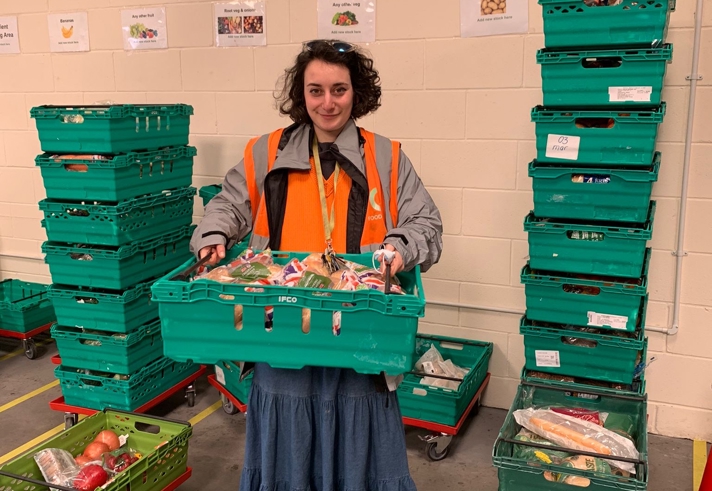 Person wearing a gray jacket and orange high-visibility vest, holding a green plastic crate filled with assorted packaged food items. The individual is standing in a warehouse-like setting with beige walls, yellow floor markings, and multiple stacks of green crates on wheeled trolleys arranged around them.