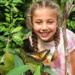 A smiling young girl with braided hair leans forward to observe a butterfly resting on a small orange flower. Surrounded by lush green tropical plants, she watches closely with curiosity. The butterfly’s patterned orange, black and white wings are in sharp focus in the foreground, creating a colourful wildlife encounter in a butterfly house or tropical garden.
