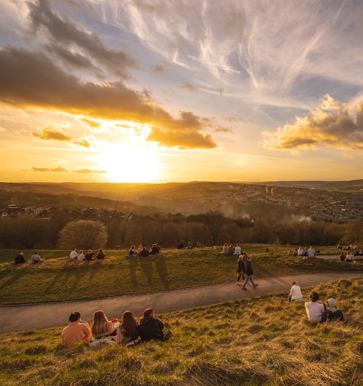 People walking along and sitting in Sheffield's Bolehills Park while they enjoy a golden sunset with views of Stannington and the Peak District beyond 