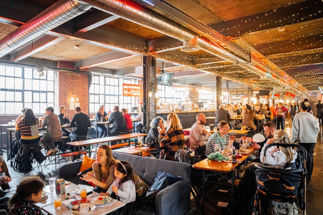 A lively indoor food hall with long wooden tables and benches filled with people eating and drinking. The space has exposed brick walls, large industrial-style windows, and string lights hanging from the ceiling, creating a warm and festive atmosphere. A stroller is visible in the foreground, and the hall extends into the background with more diners.