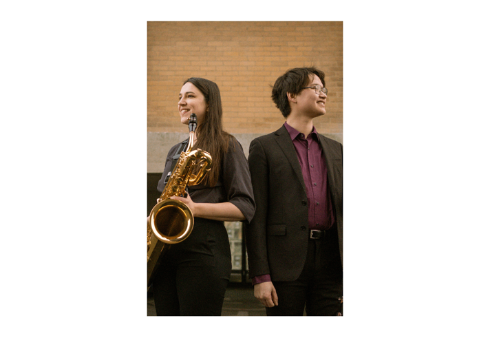 Two young musicians stand back-to-back outdoors in front of a brick building, smiling and looking in opposite directions. One holds a large saxophone and wears a dark outfit, while the other wears glasses and a dark suit with a burgundy shirt. The warm-toned portrait highlights the performers and their instruments in a relaxed, professional setting.