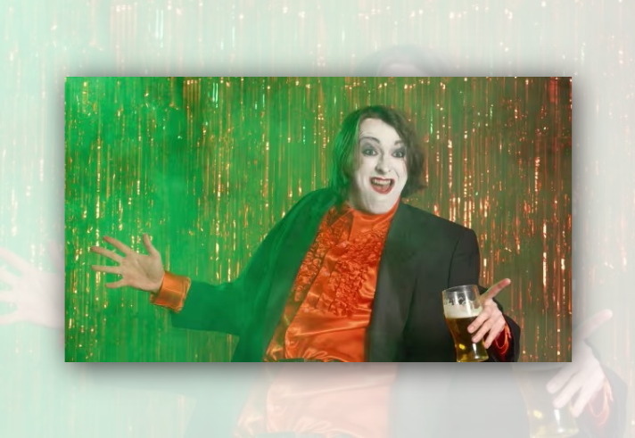 Person posing in front of a shimmering gold tinsel backdrop, lit with green stage-style lighting. Wearing a black jacket over a bright orange ruffled shirt, the person gestures with one hand while holding a pint glass in the other. The colourful, theatrical image has a cabaret or performance atmosphere, with metallic streamers filling the background.