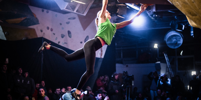 A woman  is on a indoor climbing wall. She is hanging on by a single hand and is reaching for another hand-hold. There is a large crowd  watching.