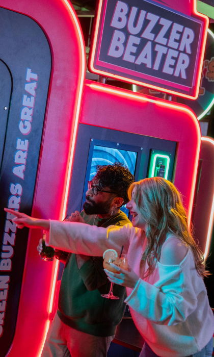 Three people playing an interactive arcade game called “Buzzer Beater” in a neon-lit venue. The game features a large vertical panel with multiple round buzzers and a digital score display reading “0550.” Bright signage on the machine includes phrases like “Green buzzers are great.” One person is actively playing, while another points at the panel, holding a drink. The setting is vibrant with colourful lights and a lively atmosphere.