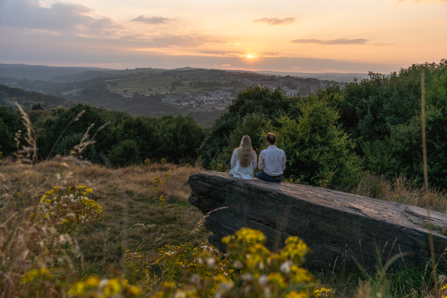 Two people sitting on a large rock in a grassy hillside area, overlooking a scenic valley with rolling green hills and a village in the distance. The sun is setting on the horizon, casting a warm golden light across the landscape, with trees and wildflowers in the foreground.