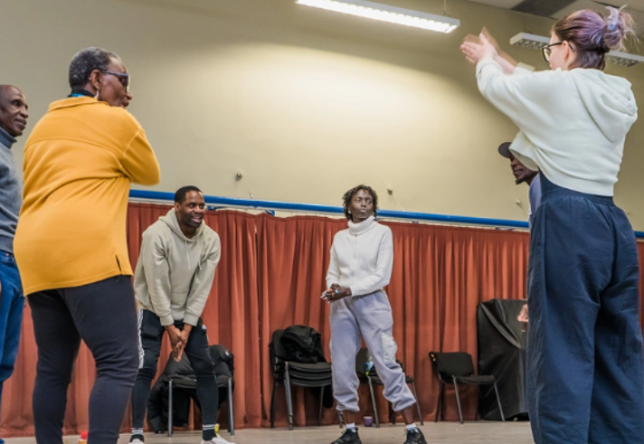 A group of six people take part in an interactive workshop or rehearsal in a community hall. Participants stand in a loose circle, with one person at the front gesturing with raised arms while others watch and respond. Chairs line the edges of the room, and red curtains hang along the back wall. The bright indoor space has cream walls, overhead strip lighting and an open floor used for movement-based activities.