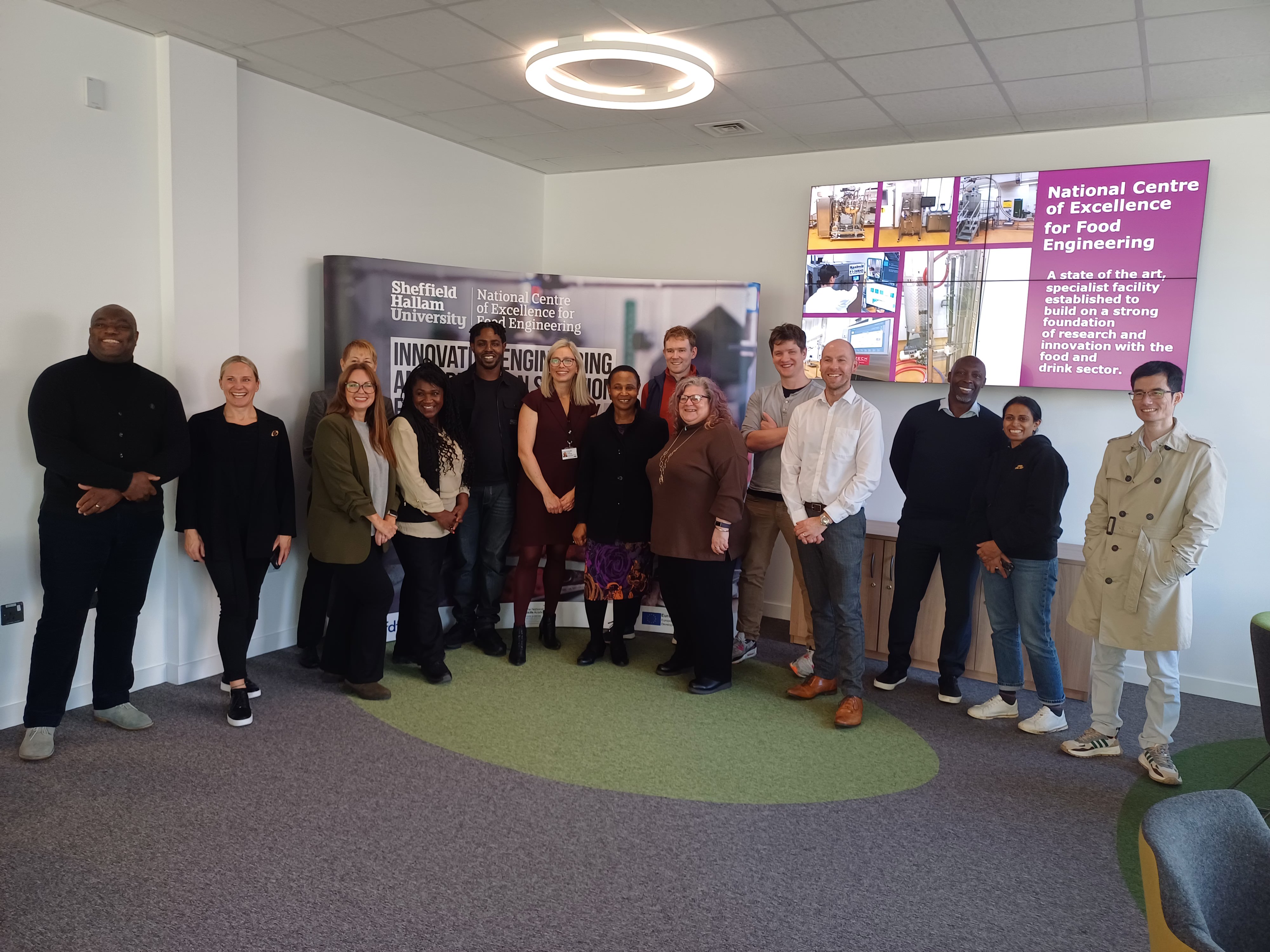 A group of twelve people standing in a semi-circle inside a modern meeting space with grey carpet and green accents. Behind them is a large banner displaying “Sheffield Hallam University” and “National Centre of Excellence for Food Engineering.” On the wall to the right, there is a digital screen showing images and text about the centre. The room has white walls, a ceiling light, and contemporary chairs arranged along the side.
