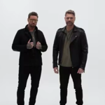 Two people stand side by side against a plain light-grey background. Both wear dark casual clothing, including jackets, slim-fit trousers and boots. One stands with hands raised near the jacket collar, while the other stands with arms relaxed by the sides. The minimalist studio-style image has no visible props, text or background details.