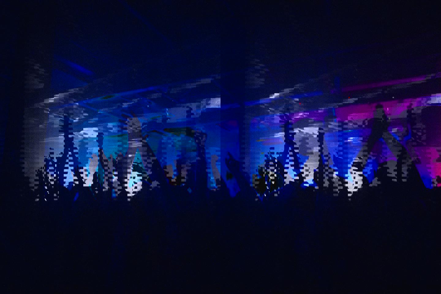 A crowd dances, arms held high, at a night club. The lighting is all blues and purples.