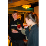 People socialising in a warmly lit bar or lounge, holding colourful cocktails. In the foreground, one person sips a pink drink from a coupe glass while another holds a red cocktail garnished with mint. A third person stands nearby with a green drink. Pendant lighting and modern décor create a relaxed evening atmosphere, with additional guests and seating visible in the background.